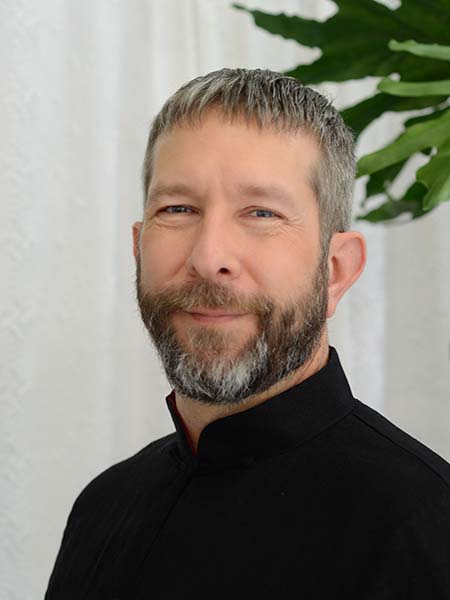 Headshot of Julian smiling. He's wearing a black banded collar and standing in front of a plant with large bright green leaves
